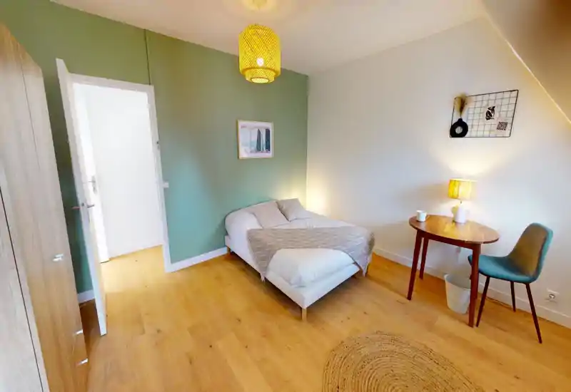 Cozy, well-lit bedroom with a neatly made double bed, soft textiles and warm wooden flooring. A small table and chair beside the bed create a practical corner for reading or working.