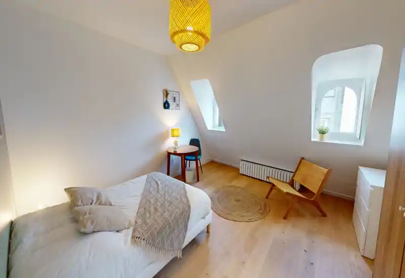 Bright, cosy bedroom with a comfortable bed, warm bedside lamp, small round table and a wicker chair by the window. Light wood floors and neutral textiles create a calm, modern atmosphere ideal for rental listings.