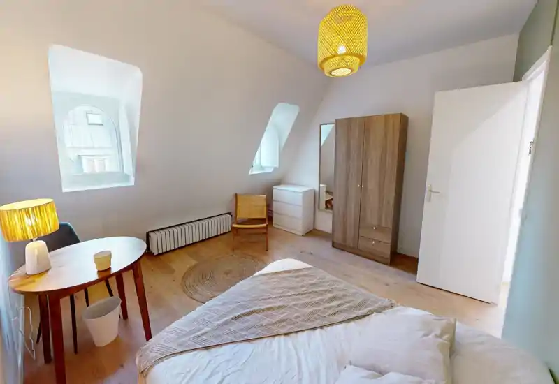 Bright, minimalist bedroom with a neatly made bed in the foreground, two angled windows providing natural light, a wooden wardrobe and simple rattan accents for a cozy, Scandinavian feel.
