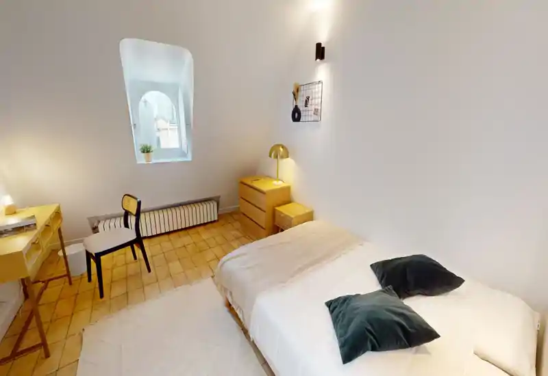 Chambre lumineuse et minimaliste avec un lit confortable, coussins moelleux et mobilier en bois chaleureux. Un petit bureau et une chaise près de la fenêtre en arc la rendent pratique pour travailler ou lire.