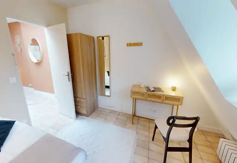 Coin chambre lumineux et chaleureux avec lit visible, petit bureau et chaise, armoire et miroir mural — éclairage doux et décoration neutre pour une ambiance accueillante.