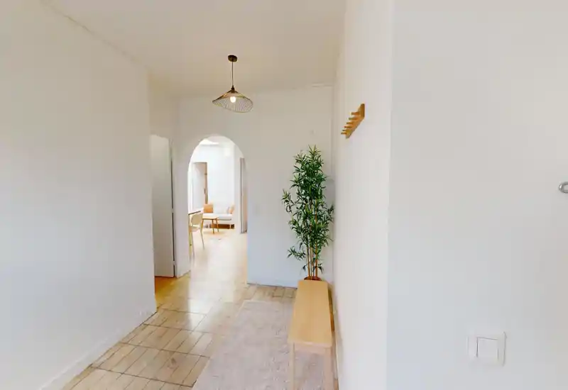 Bright, minimal entrance hallway with a bench, rug and wall hooks leading into a living area through an arched doorway.