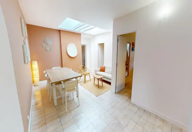 Bright, modern dining area with a wooden table and several chairs, adjacent small lounge with a sofa and rattan chair; warm accent wall and skylight create an inviting atmosphere.