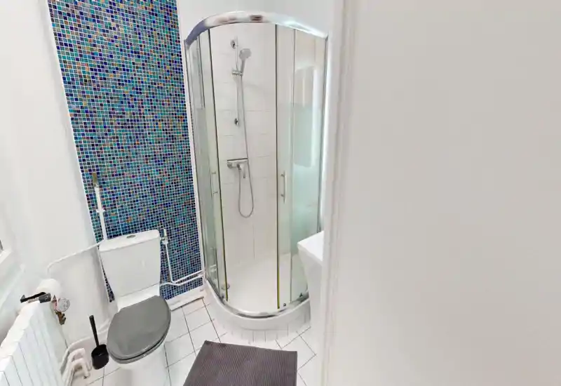 Compact modern bathroom with corner glass shower, white tiles and a toilet; blue mosaic accent wall adds character.