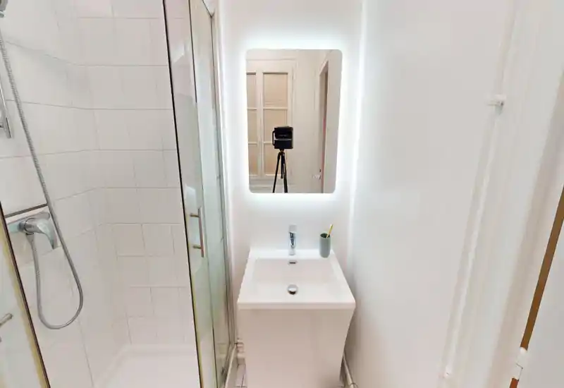 Compact modern bathroom with a tiled shower, glass door and a small white sink under a backlit mirror — clean, minimal and well lit.