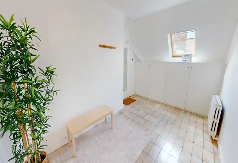 Bright, minimal entryway with a small bench, coat hook and built-in storage under a skylight — clean tiled floor and a radiator make it practical and welcoming for guests.