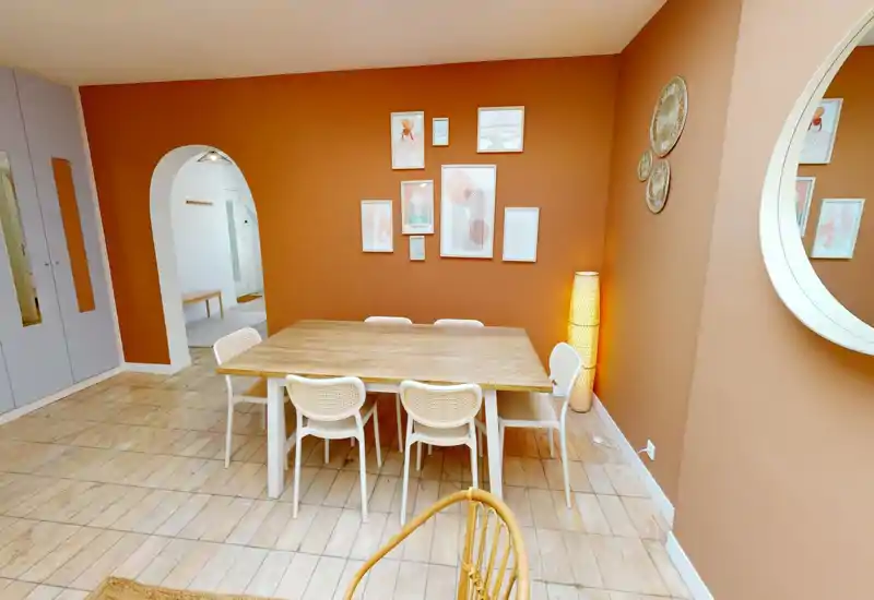 Spacious, styled dining room with a large wooden table and six woven chairs against a warm terracotta wall, accented by framed art and a standing lamp for a cozy, inviting atmosphere.