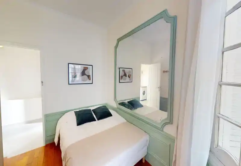 Bright, cozy bedroom with a double bed dressed in neutral linens, two dark accent pillows and decorative wall paneling. A large framed mirror and window provide ample light and a sense of space.