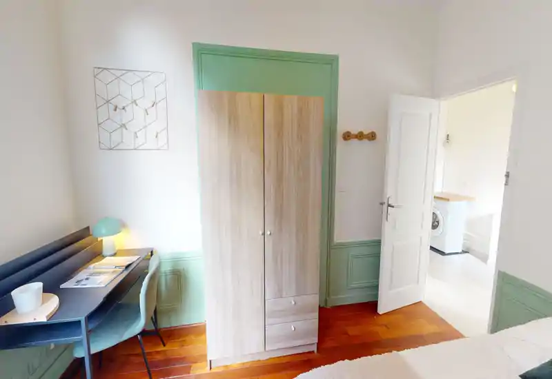 Compact, well-lit bedroom corner with a visible wardrobe, small desk and a portion of the bed. Soft green trim, warm wooden floor and a bedside lamp give the room a fresh, welcoming feel — ideal for single occupancy or a short-stay.