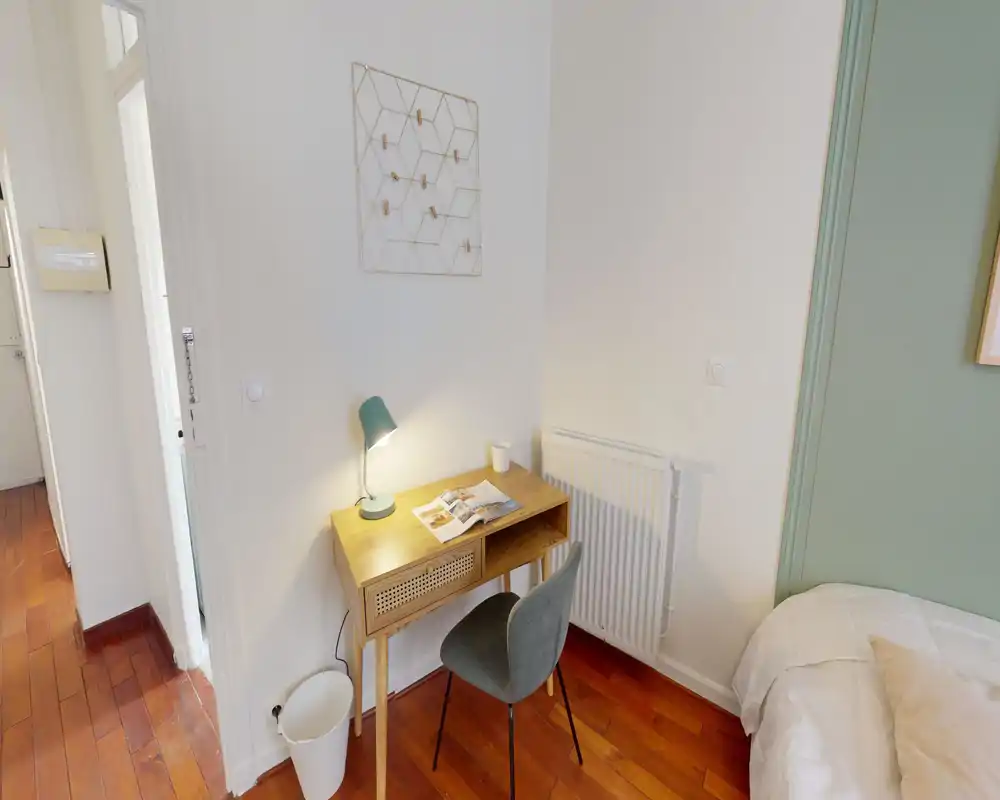 Angolo camera compatto con una piccola scrivania in legno, lampada e un letto matrimoniale parzialmente visibile; il parquet caldo e l'arredo dai toni neutri creano un ambiente accogliente con zona studio.