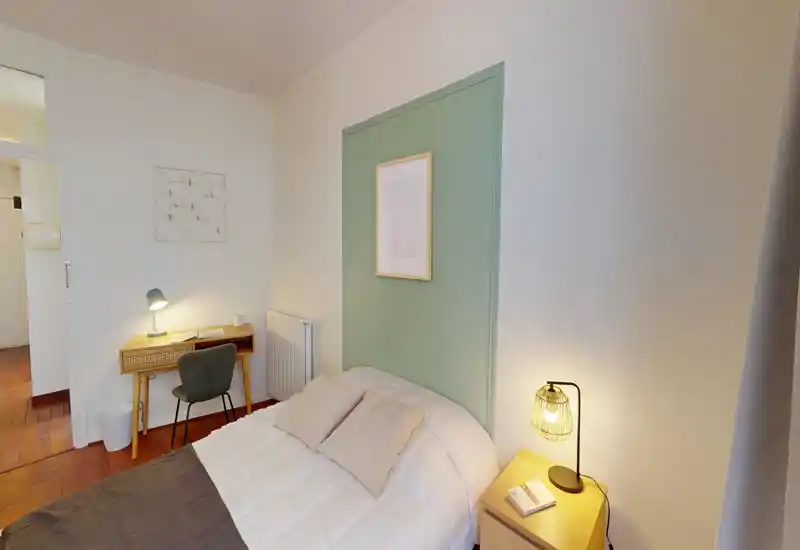 Camera singola accogliente e ben illuminata con letto confortevole, lampada da comodino e una piccola scrivania — pannello verde a contrasto e pavimento in legno caldo.