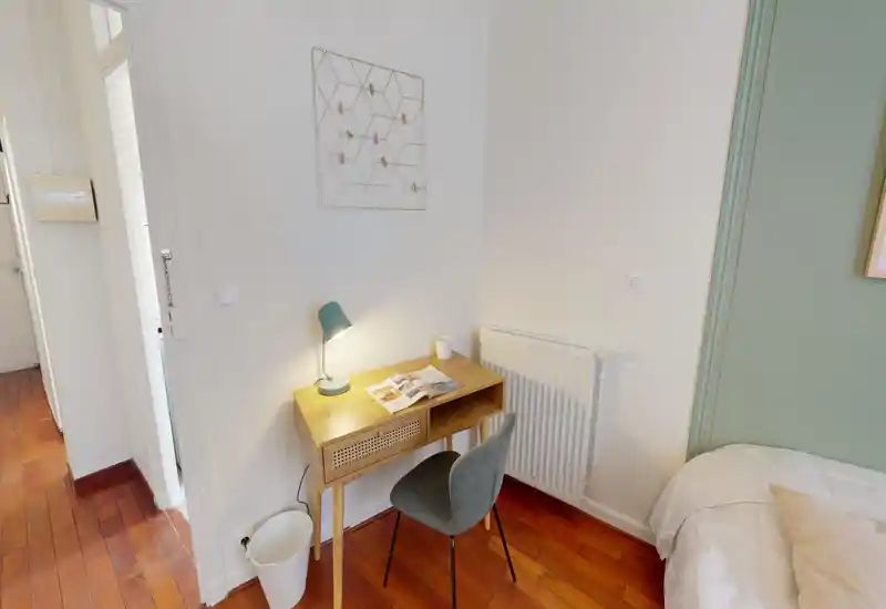 Angolo camera compatto con una piccola scrivania in legno, lampada e un letto matrimoniale parzialmente visibile; il parquet caldo e l'arredo dai toni neutri creano un ambiente accogliente con zona studio.