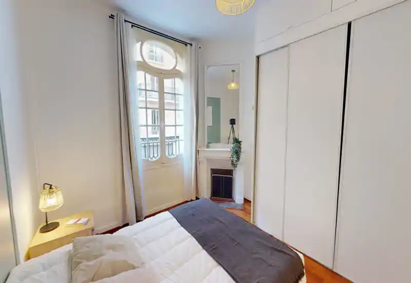 Camera da letto luminosa e ben arredata con un letto confortevole, lampada da comodino e una grande finestra che illumina la stanza; armadi a muro e uno specchio decorativo completano l'ambiente.