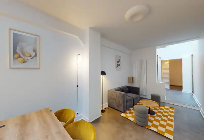 Salon moderne et lumineux avec un canapé gris compact, tapis jaune et blanc à carreaux, poufs et éclairage chaleureux — un espace confortable et bien décoré pour se détendre.