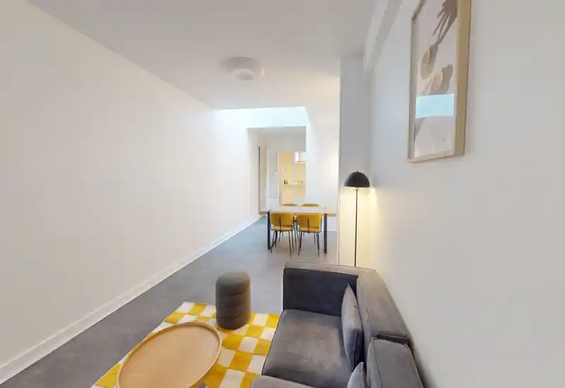 Espace séjour moderne et minimaliste avec un petit canapé, un tapis à motifs et une table basse; une table à manger avec chaises jaunes est visible en arrière-plan dans une pièce lumineuse.