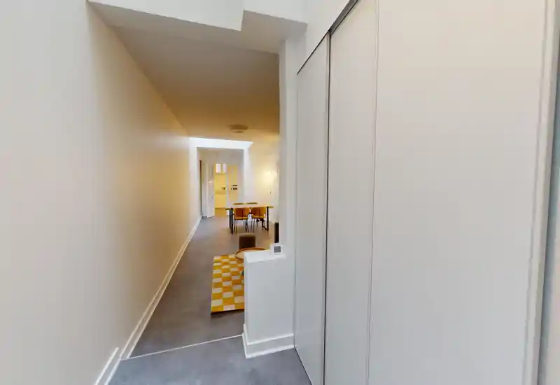 Vue en longueur minimaliste d'un salon/couloir menant à une salle à manger lumineuse ; décoration épurée avec un tapis à carreaux et des chaises modernes.
