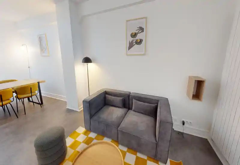 Espace de vie élégant avec un petit canapé gris, tapis à motifs et lampe sur pied chaleureuse ; espace ouvert donnant sur une table à manger avec chaises jaunes — lumineux, moderne et bien mis en scène pour la location.