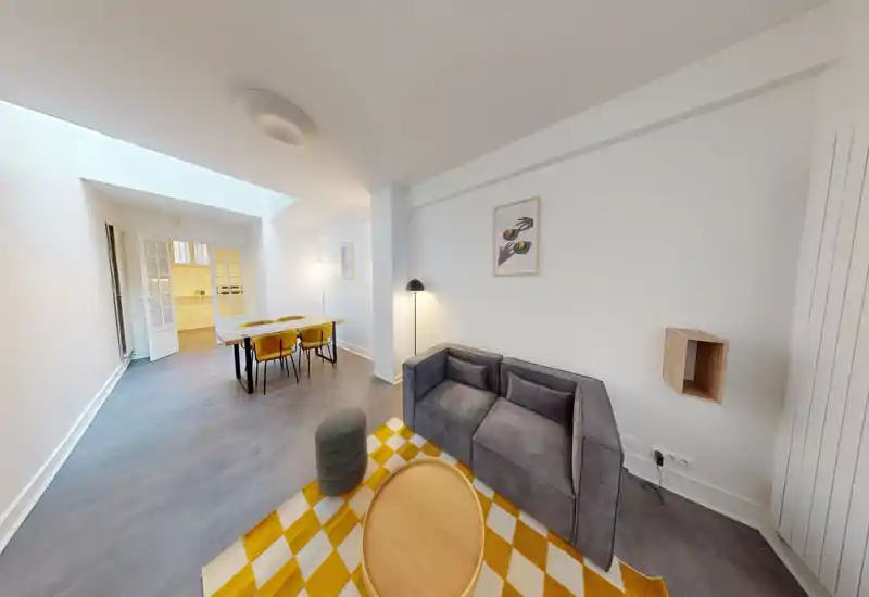 Bright, modern living area with a compact gray sofa, patterned yellow rug and a small coffee table; a dining table with chairs sits further back next to an open doorway leading to the kitchen. Clean white walls, soft lighting and contemporary accents make this a welcoming communal space.