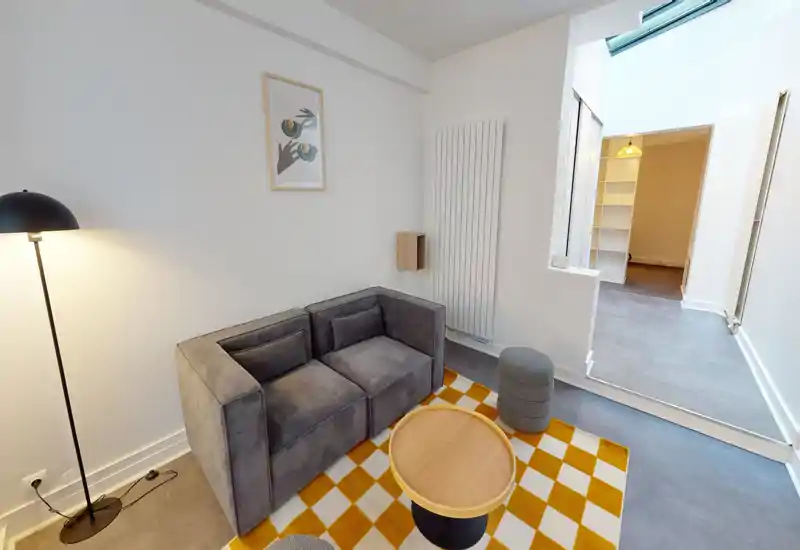 Petite pièce à vivre avec canapé gris deux places, tapis à motif et table basse ronde ; lampe sur pied et lumière naturelle de la pièce adjacente la rendent chaleureuse.