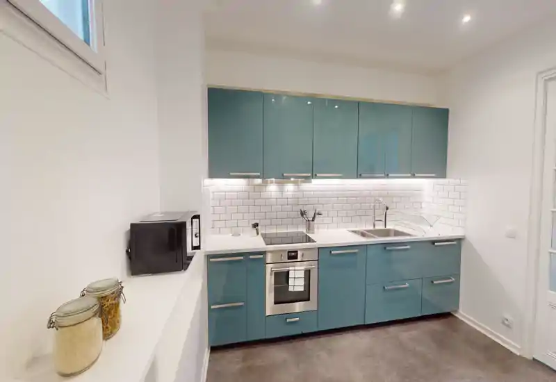 Cuisine moderne et compacte avec placards laqués bleu-vert, four encastré, plaque à induction et évier en inox ; éclairage sous les meubles et plan de travail propre, idéale pour un petit appartement.