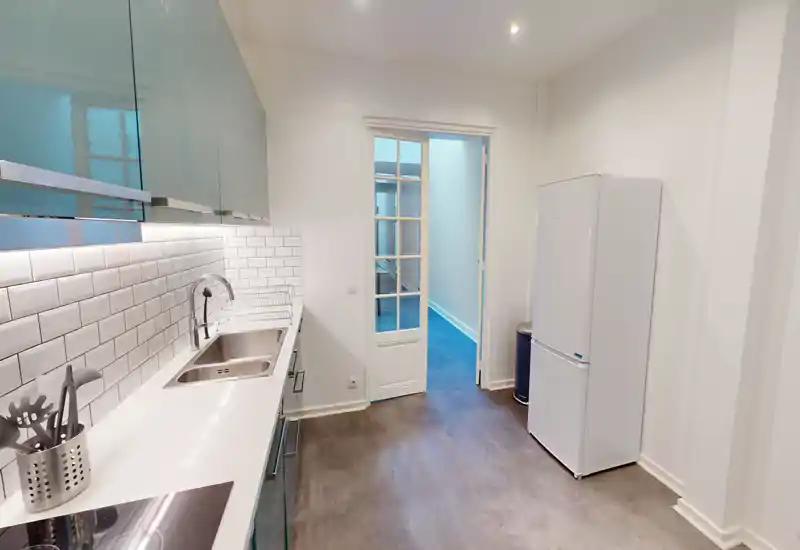 Cuisine moderne et étroite avec plan de travail blanc, crédence en carreaux métro, évier intégré et réfrigérateur indépendant ; lumineuse, propre et bien agencée pour un appartement urbain.