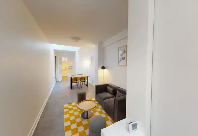 Coin salon moderne et compact avec un canapé deux places sombre, tapis damier jaune et petite table basse, s'ouvrant sur la salle à manger et la cuisine. Espace lumineux et soigné, parfait pour une annonce en centre-ville.