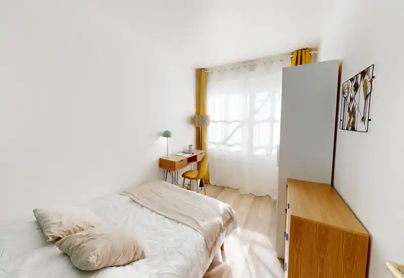 Chambre lumineuse et minimaliste avec un lit bien fait, mobilier en bois chaleureux et un petit bureau près de la fenêtre. Voilages et touches moutarde créent une ambiance cosy et lumineuse, idéale pour la location courte durée.