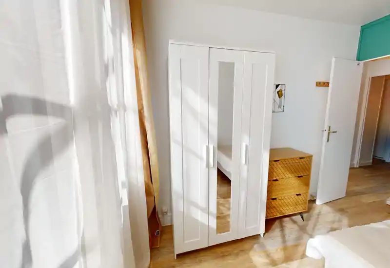 Chambre lumineuse et sobrement meublée avec un coin lit visible, une armoire blanche à porte miroir et une commode en bois; la lumière naturelle chaude baigne la pièce.
