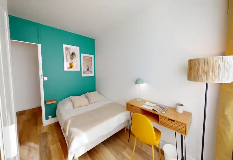 Chambre compacte et lumineuse avec un lit, un mur d'accent turquoise et un petit bureau en bois avec chaise jaune — bien éclairée et mise en valeur pour une personne.