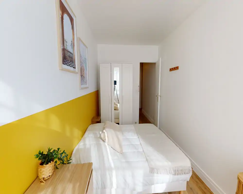 Camera da letto luminosa e moderna con letto bianco trapuntato, parete d'accento gialla e mobili in legno chiaro. La stanza è ariosa e curata con una pianta e quadri, perfetta per un annuncio in affitto.