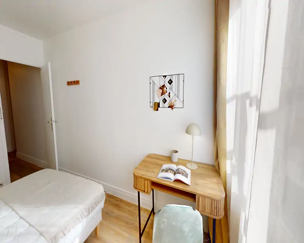 Angolo camera da letto luminoso e minimale con letto parzialmente visibile, scrivania in legno con lampada e libro aperto, e luce naturale morbida dalla finestra con tenda.