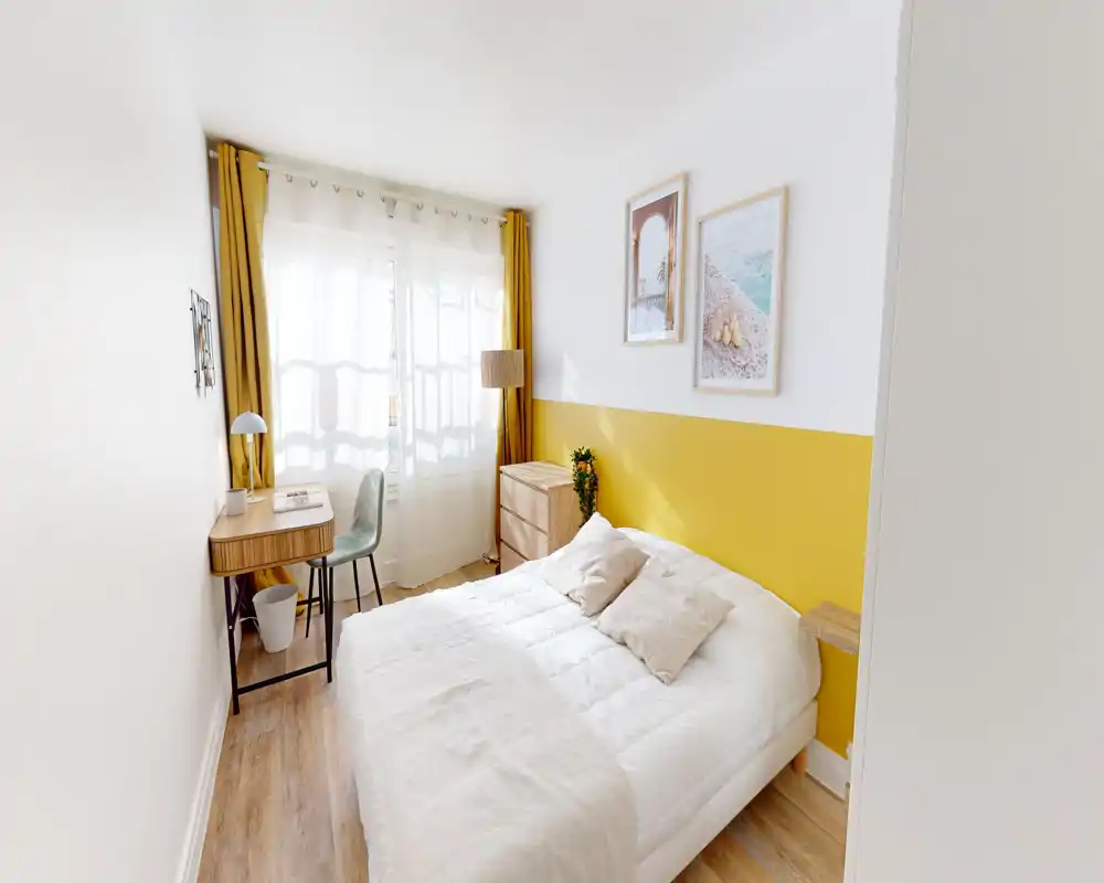 Camera da letto luminosa e compatta con un letto matrimoniale ben rifatto, biancheria neutra e una parete d'accento gialla. Una piccola scrivania vicino alla finestra e la luce naturale la rendono adatta per lavoro e riposo.