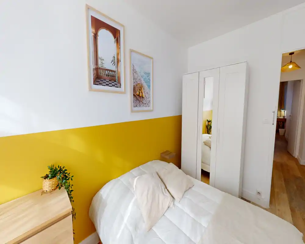 Camera da letto luminosa e curata con armadio bianco e letto matrimoniale accogliente contro una parete accentata gialla. Luce naturale e tessili morbidi creano un'atmosfera invitante, perfetta per gli annunci di affitto.
