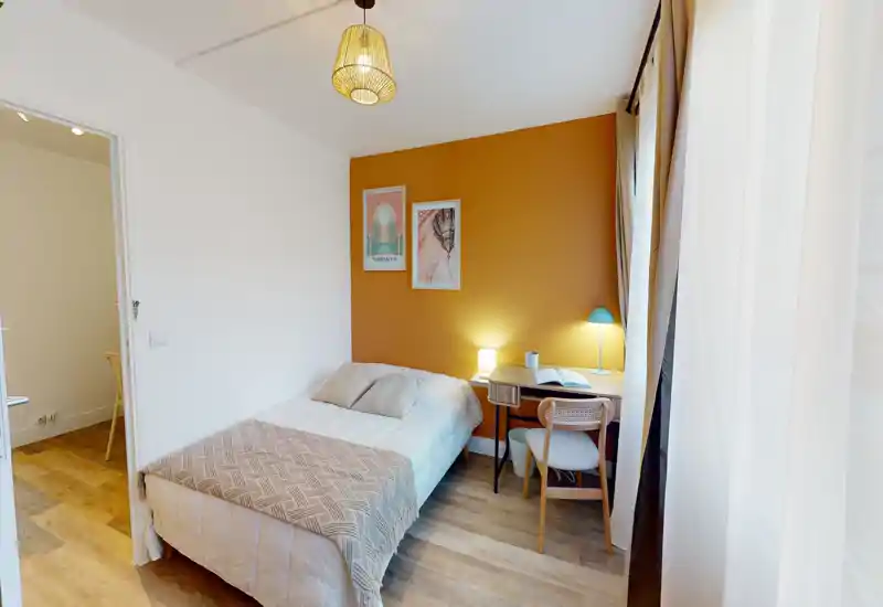 Camera singola compatta e ben arredata con parete d'accento color senape, letto singolo confortevole, lampade da comodino e una piccola scrivania vicino alla finestra — luminosa e accogliente.
