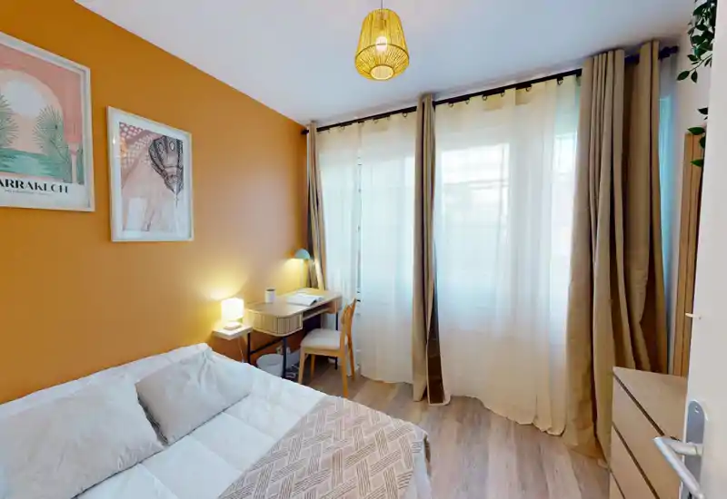 Camera da letto luminosa e arredata con letto matrimoniale, lampada da comodino e piccolo scrittoio vicino alle ampie finestre con tende; parete accentuata color ocra e tessili morbidi rendono l'ambiente accogliente.