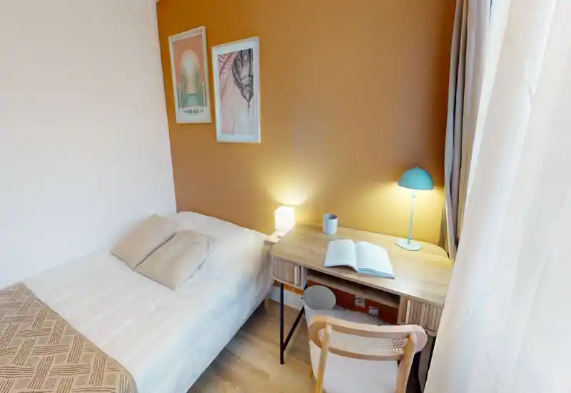 Piccola camera da letto accogliente con letto singolo sistemato, parete in tonalità calda e scrivania in legno con lampada da lettura vicino alla finestra — luminosa e ben arredata, perfetta per studenti o brevi soggiorni.