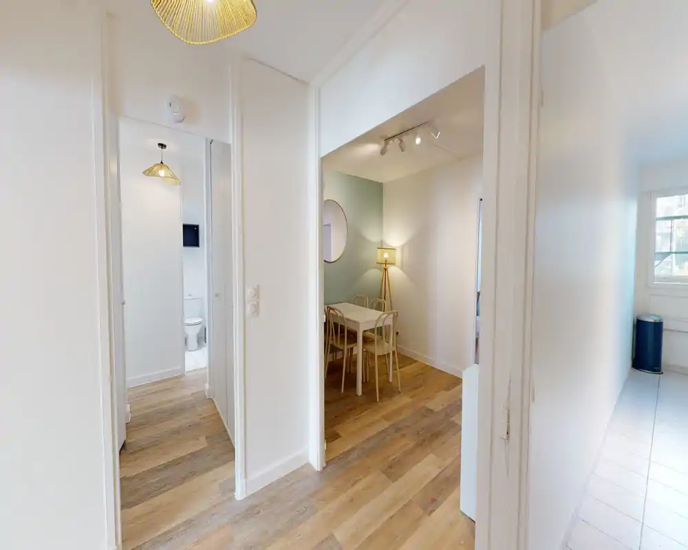 Entrée lumineuse et moderne s'ouvrant sur un espace salon/salle à manger avec une petite table, des chaises et une lampe chaleureuse — idéal pour annonces d'appartements compacts.