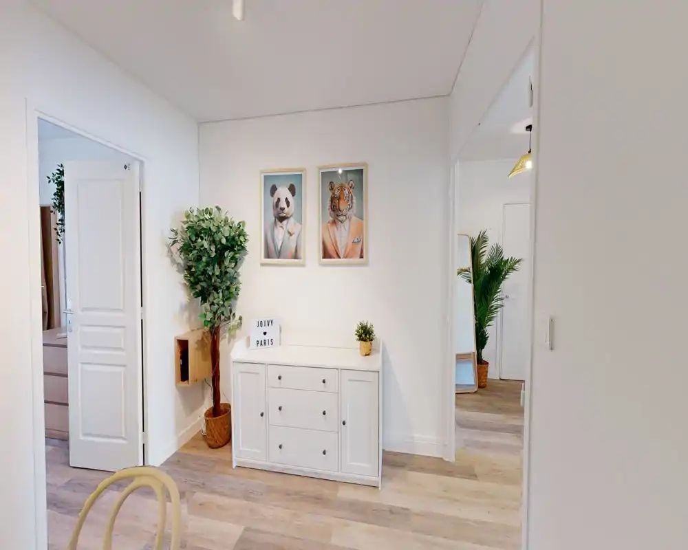 Entrée lumineuse et minimaliste avec un buffet blanc, des cadres décoratifs et des plantes. Parquet clair et murs neutres pour un espace d'accueil aéré et accueillant.