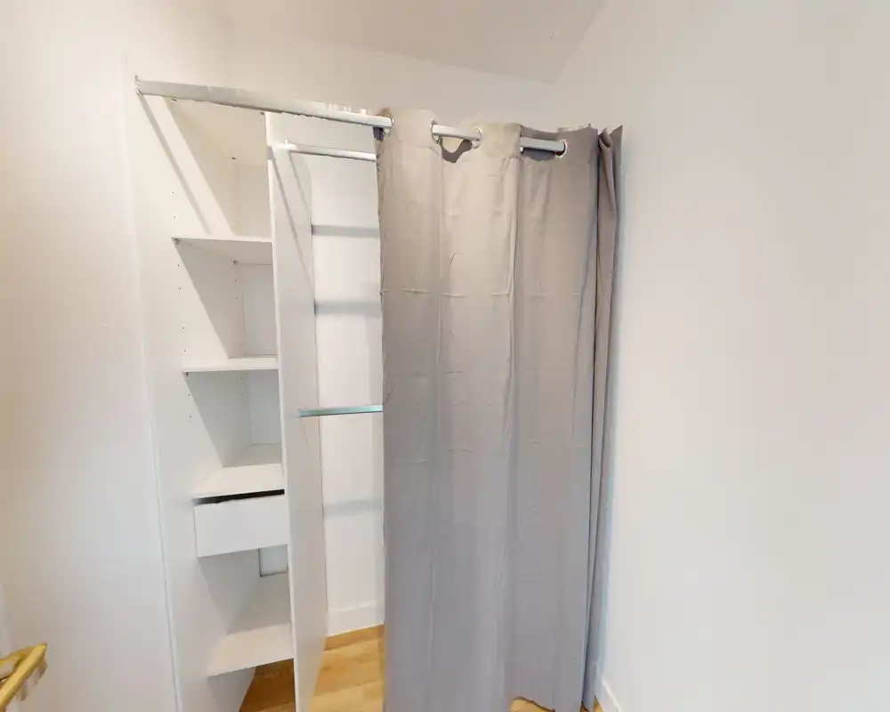 Petit dressing avec étagères blanches intégrées et rideau gris ; sol en bois clair et murs blancs créent un espace de rangement minimal et propre.