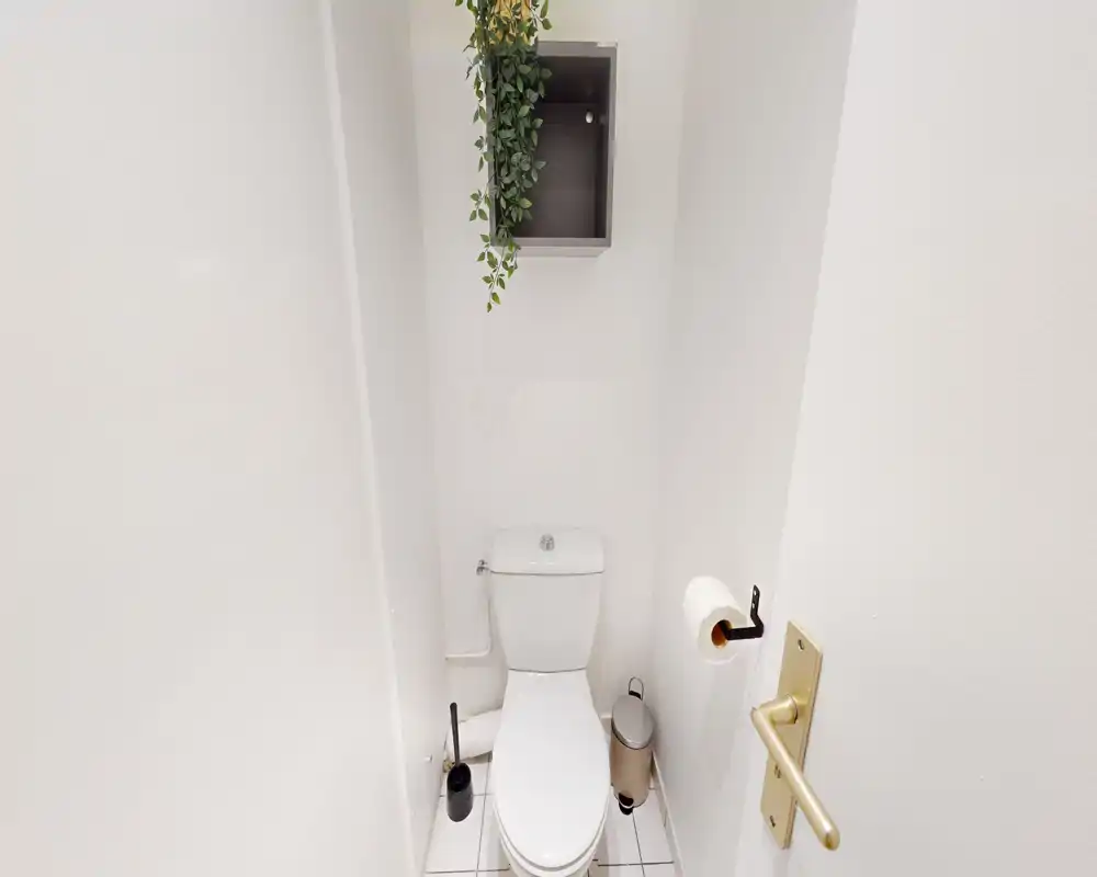 Toilettes compactes et propres avec carrelage blanc et réservoir simple ; une petite étagère végétale apporte une touche de fraîcheur.