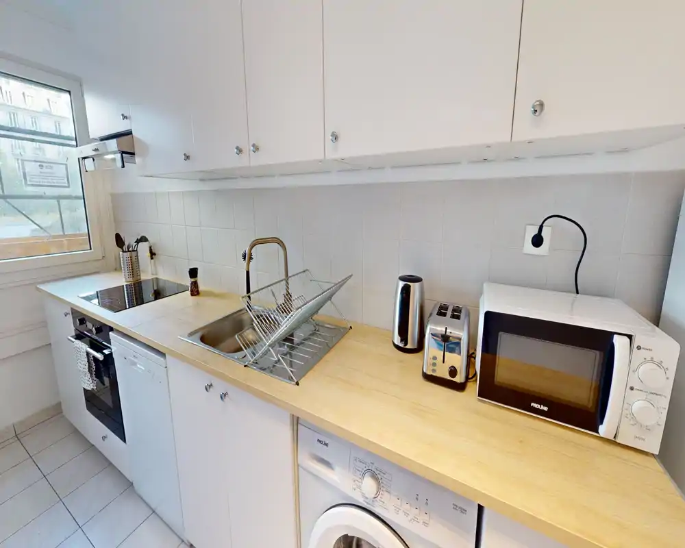 Cuisine compacte et lumineuse avec plan de travail en bois, évier avec égouttoir, plaque de cuisson et appareils intégrés (four, lave-linge, grille-pain et micro-ondes) — fonctionnelle et bien rangée.