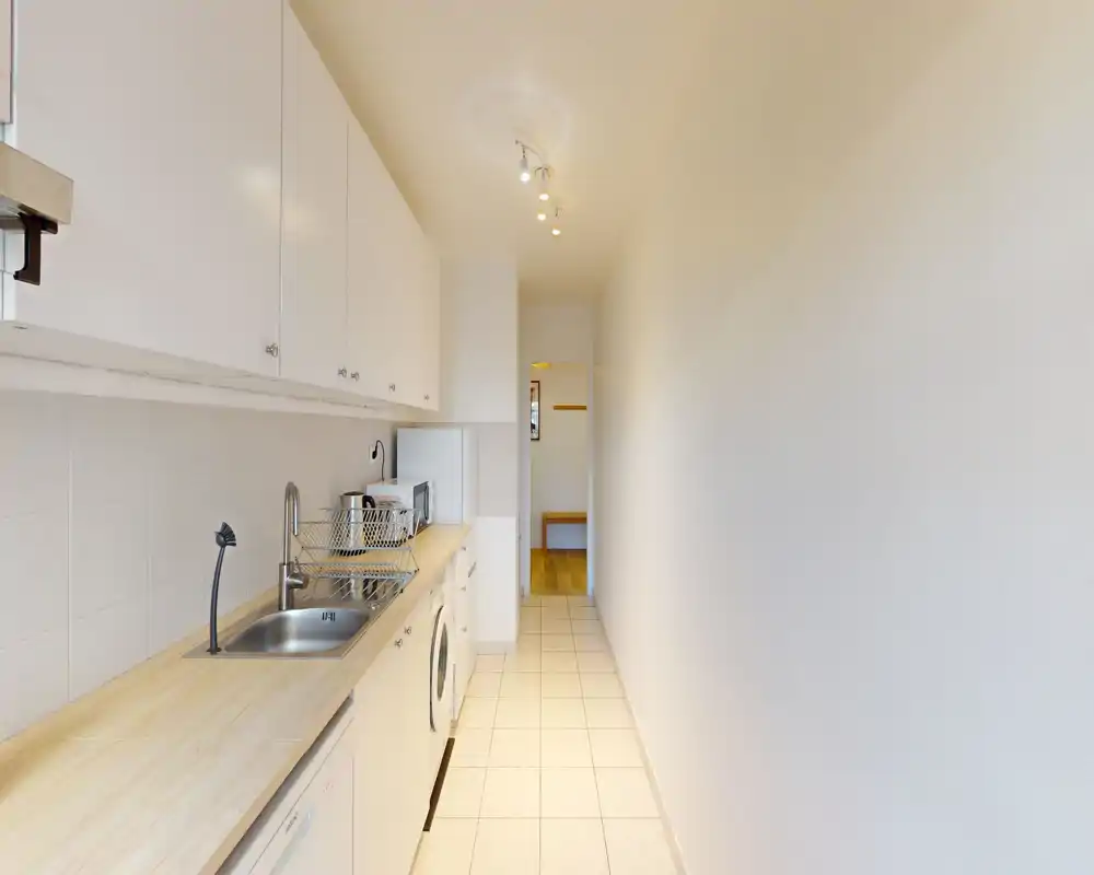 Cuisine en enfilade étroite avec plans de travail en bois clair, évier intégré, lave-linge et placards hauts ; agencement lumineux et fonctionnel adapté aux petits appartements.