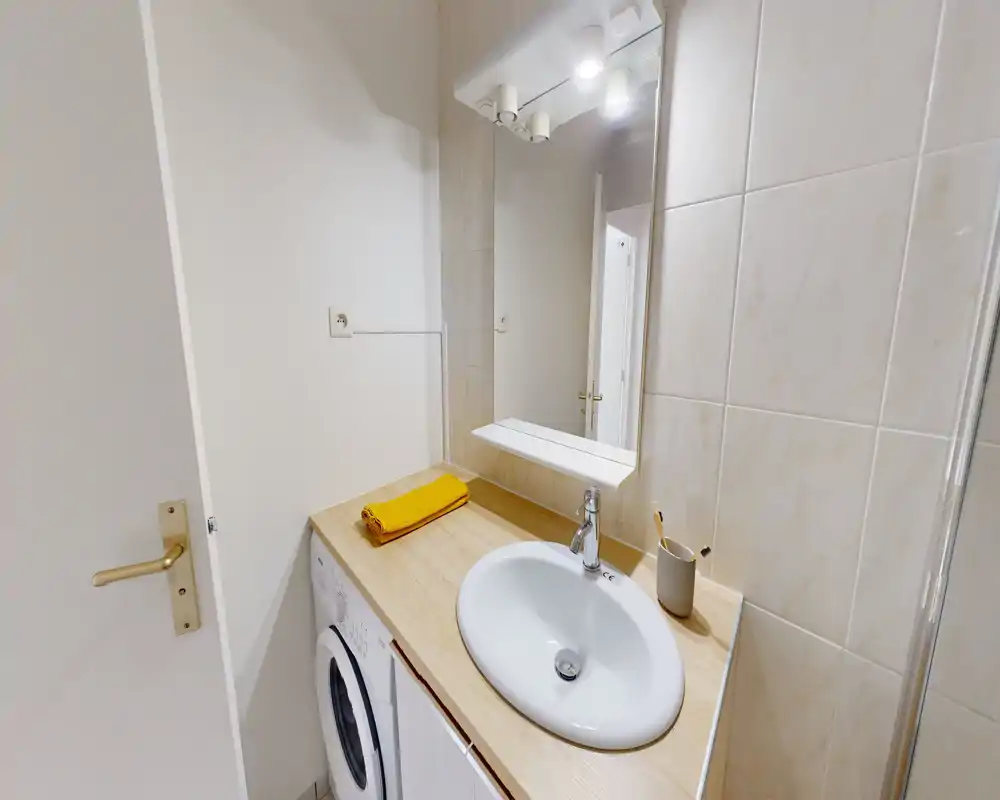 Salle de bains compacte avec lavabo rond intégré dans un plan de travail en bois, miroir mural éclairé et lave-linge sous le plan; carreaux neutres et accents chauds pour un espace propre et fonctionnel.