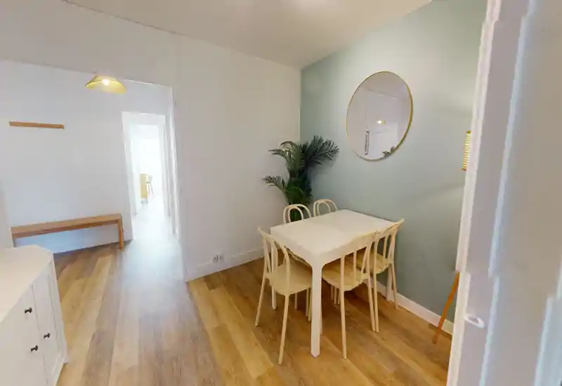 Cozy dining nook with a small white table and four matching chairs, soft pastel wall, round mirror and a decorative plant — bright and welcoming for meals.