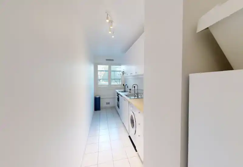 Narrow galley kitchen with white cabinets, wooden countertop, integrated appliances and a window providing natural light — compact and functional layout.