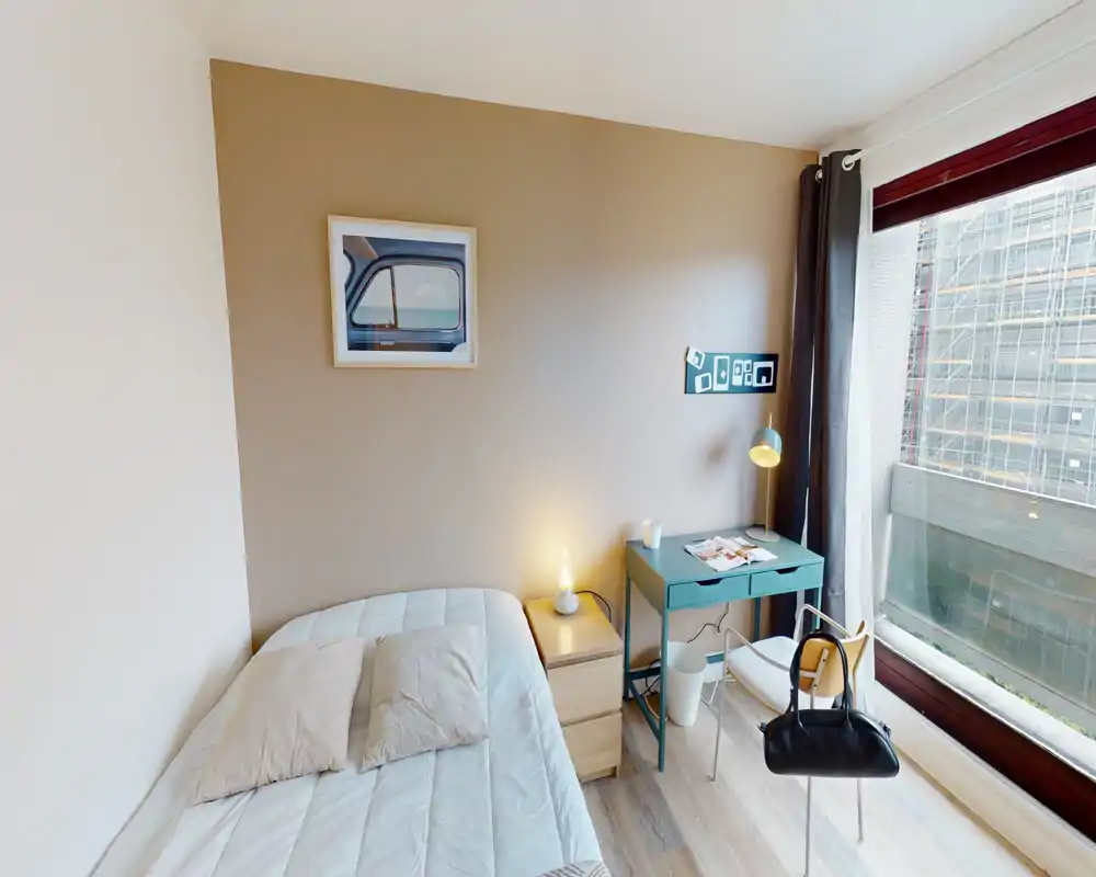 Chambre lumineuse et compacte avec un lit simple, une table de chevet et un petit bureau près d'une grande fenêtre qui laisse entrer beaucoup de lumière naturelle.