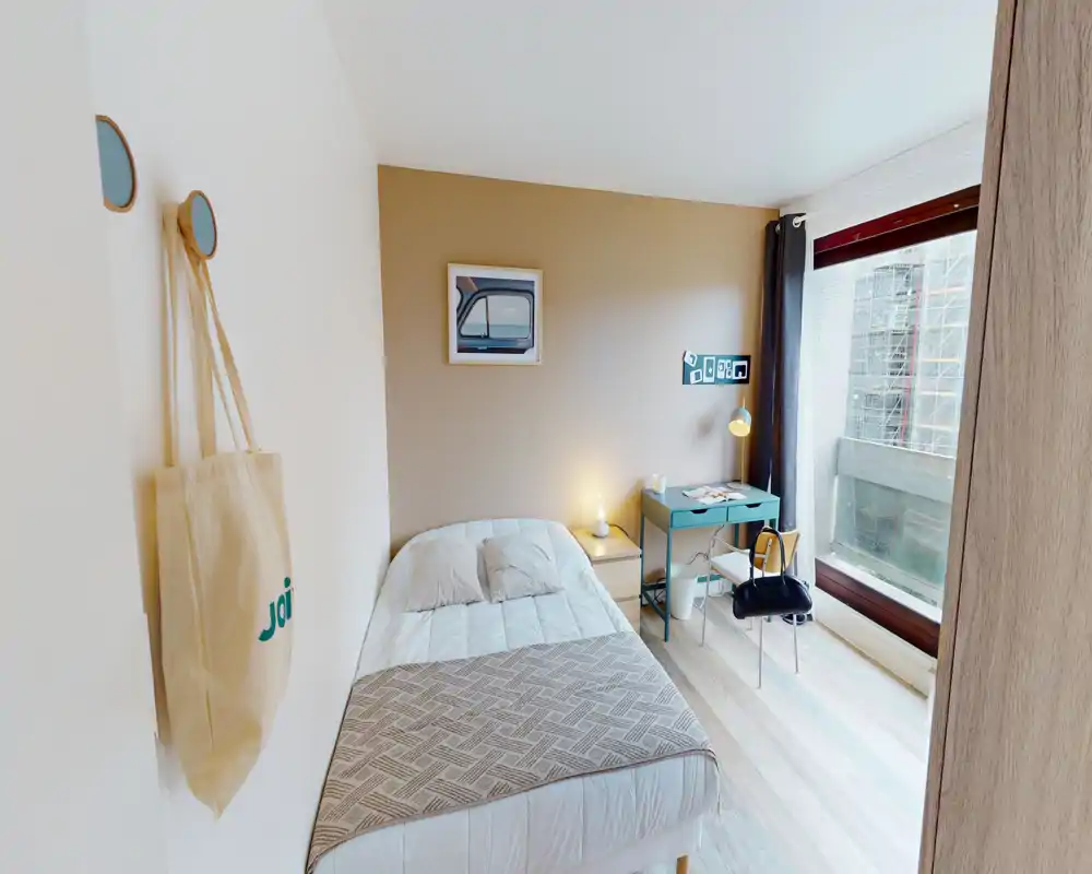 Chambre simple lumineuse et bien mise en scène avec un lit confortable, une table de chevet et un petit bureau près d'une grande fenêtre laissant entrer la lumière naturelle.
