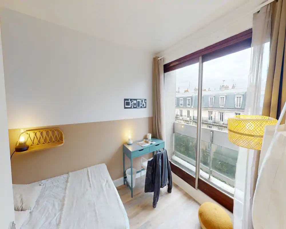 Chambre compacte et lumineuse avec un lit confortable, une lampe de chevet et un petit bureau près de la fenêtre avec vue sur la ville — style chaleureux et moderne idéal pour une personne seule.