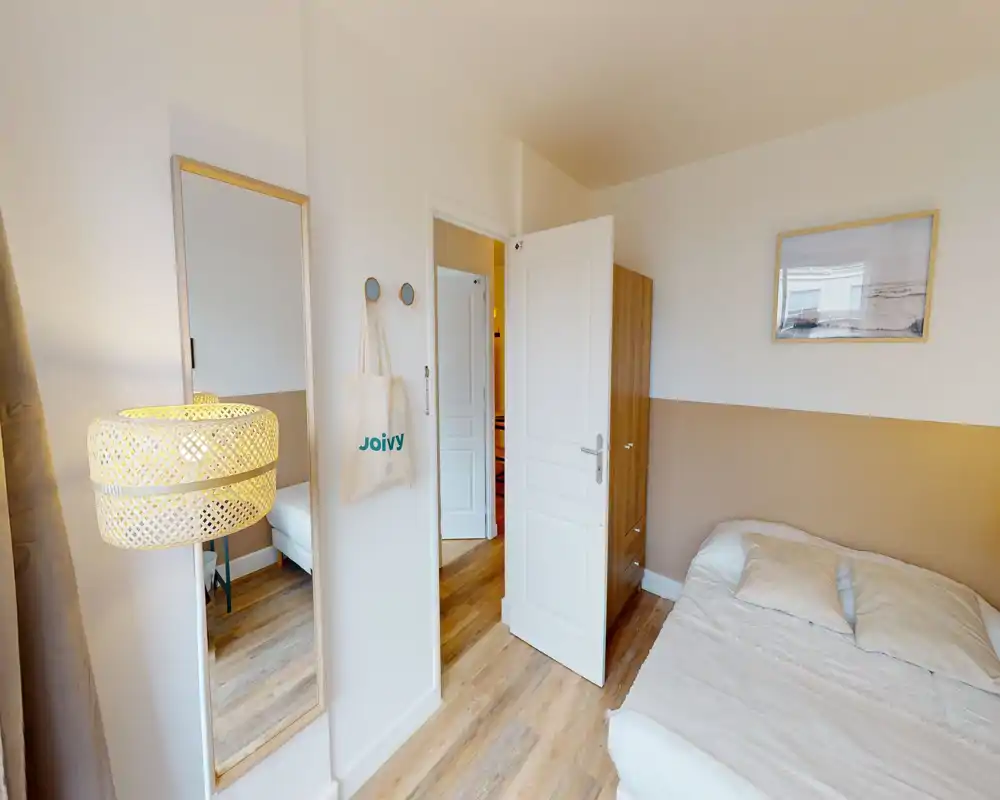 Chambre à coucher lumineuse et cosy avec lit double, parquet et décoration aux tons neutres. Un miroir sur pied et une lampe douce apportent de la profondeur et de la chaleur.