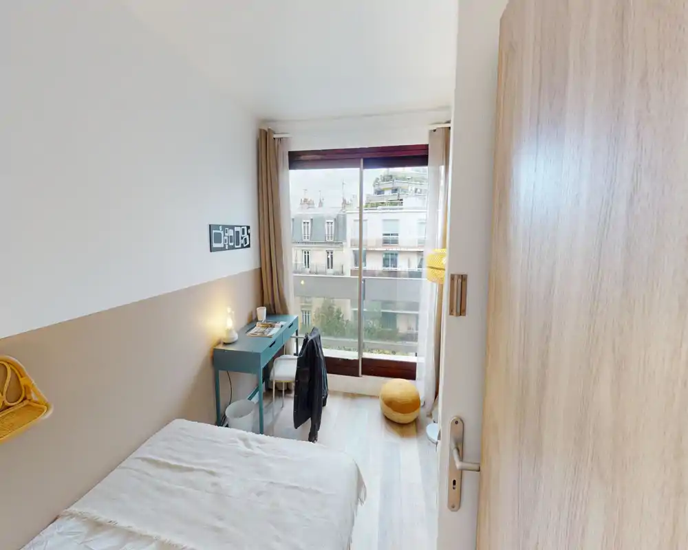 Chambre simple moderne et lumineuse avec lit confortable, bureau près de la fenêtre et grandes baies vitrées laissant entrer la lumière naturelle et offrant une vue sur la rue — idéale pour étudiant ou professionnel.